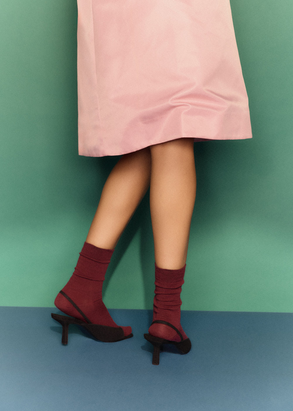 A person in a pale pink skirt stands on a blue floor against a green background, wearing Dark Plum Merino Socks with black heeled sandals. The odour-resistant calf-length socks complement their bare legs as they cross their feet.