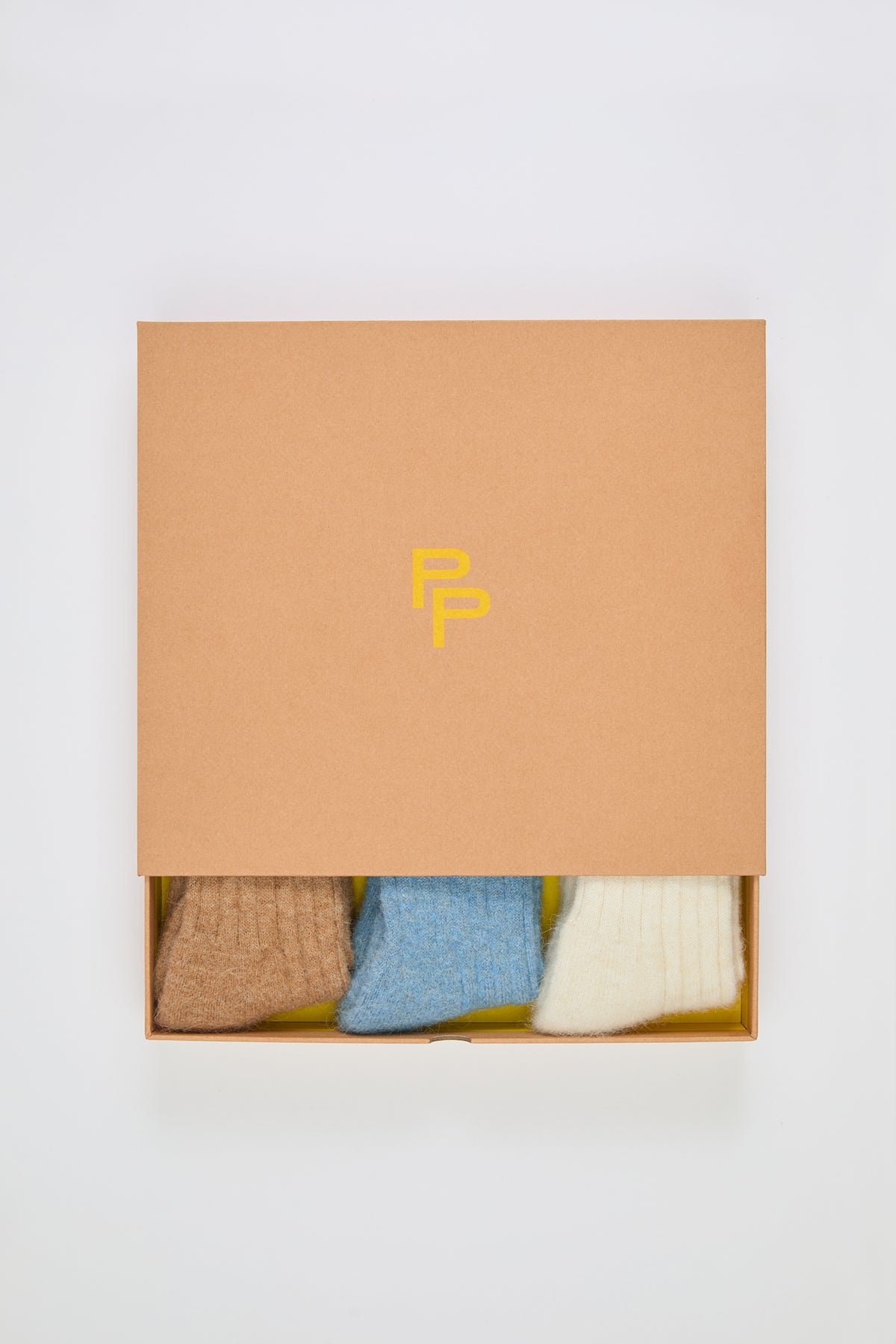 A brown box with a yellow PP logo is partially open, revealing The Ultra Soft Alpaca Ribbed Gift Set One, three neatly folded ultra soft alpaca bed socks, against a yellow interior.