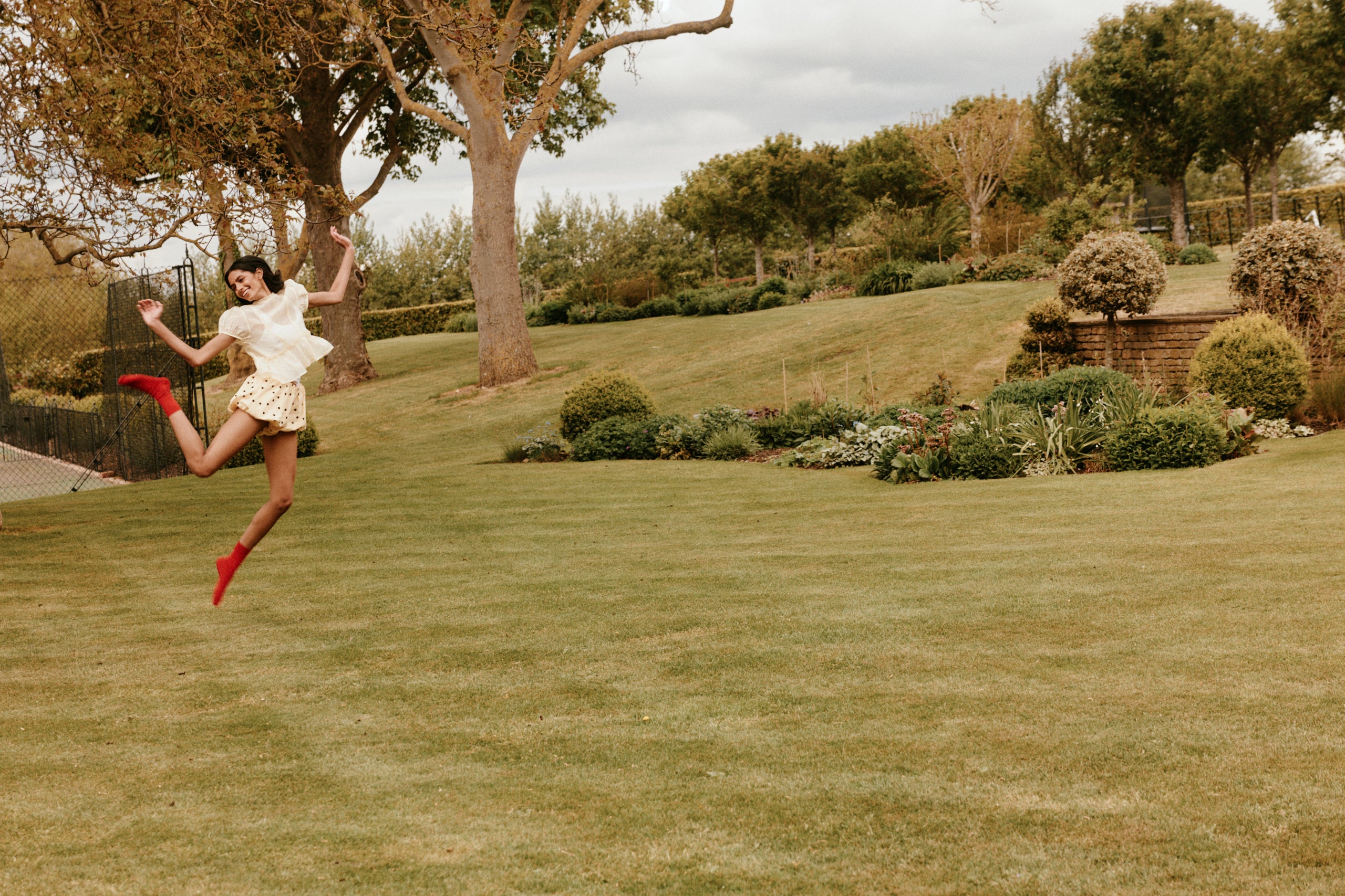 Woman in a white dress and red shoes jumping in a garden with trees and shrubs.