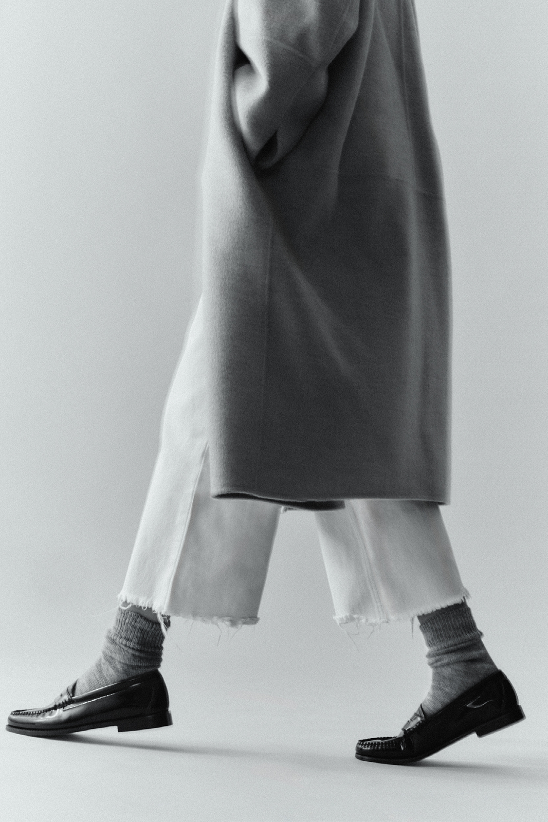 Person wearing a long coat, white trousers, grey alpaca calf length socks and black shoes on a plain background