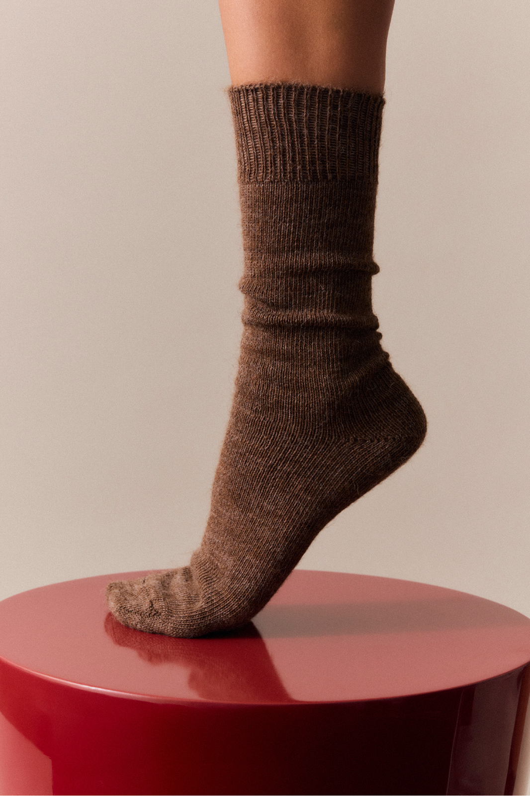 Leg in a mid-brown alpaca calf length sock on a red surface with a neutral background