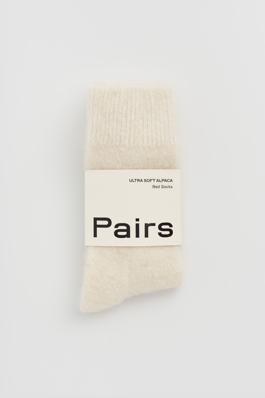 Pair of cream ultra soft alpaca bed socks with a 'Pairs' packaging band on a light grey background