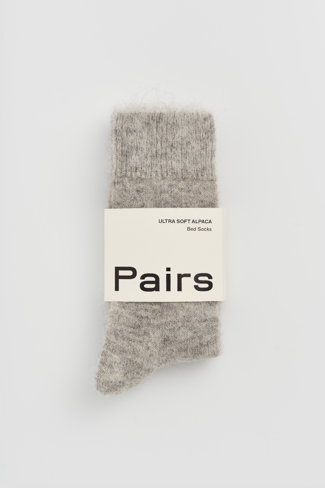 Pair of grey ultra soft alpaca bed socks with a 'Pairs' packaging band on a light grey background