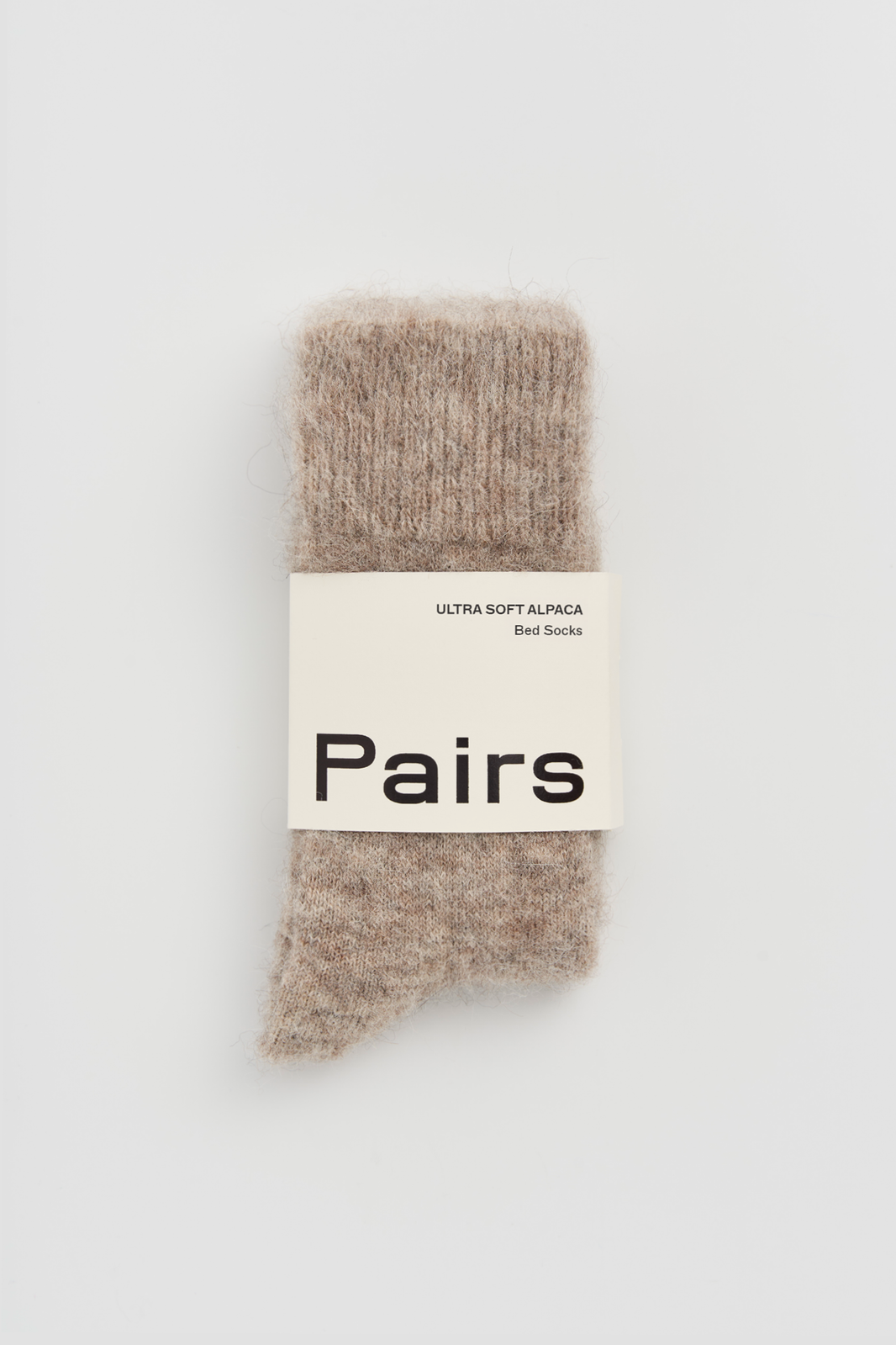 Pair of oatmeal ultra soft alpaca bed socks with a 'Pairs' packaging band on a light grey background